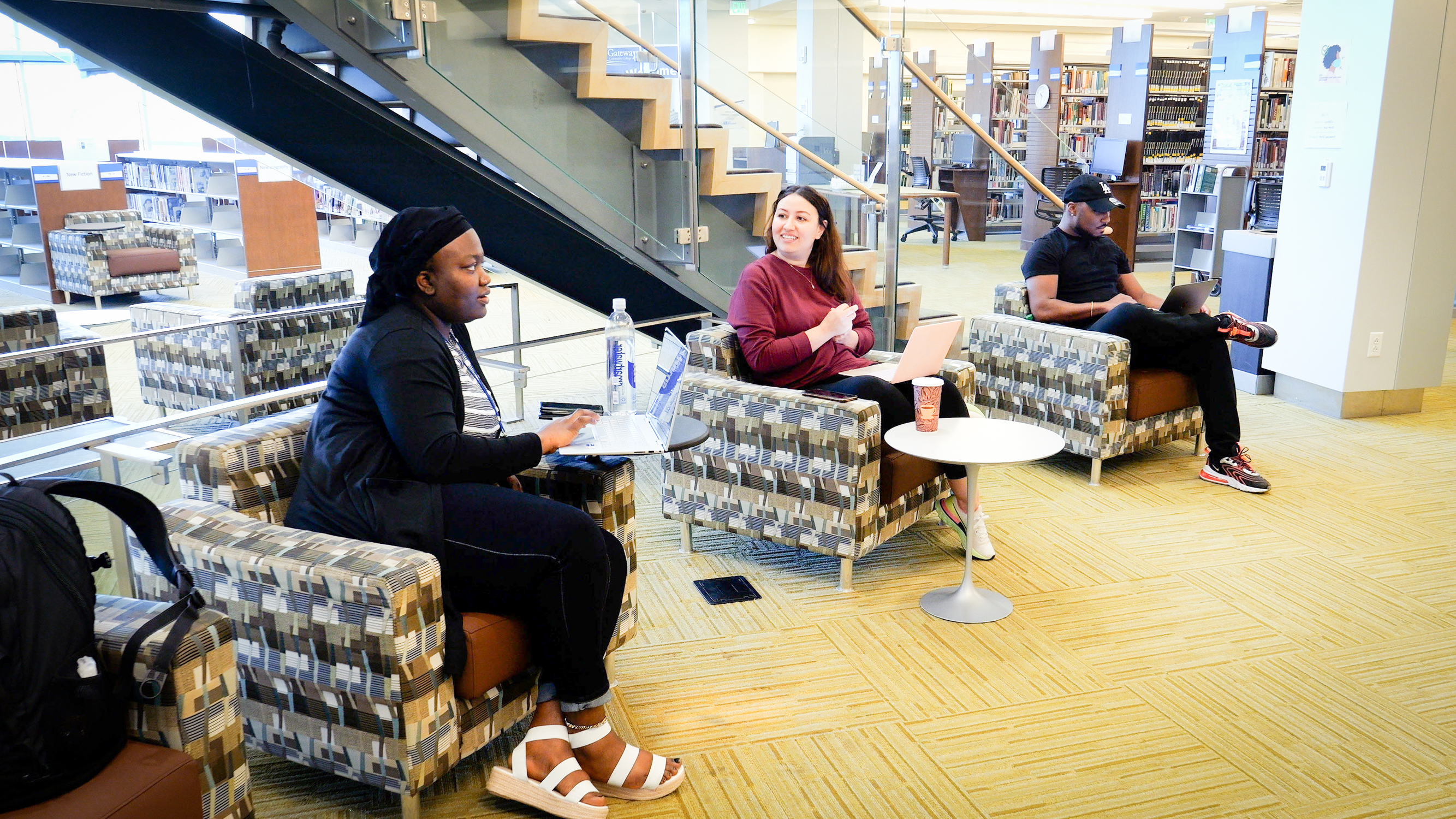 Three students seated in chairs, engaged in reading within a quiet library setting.